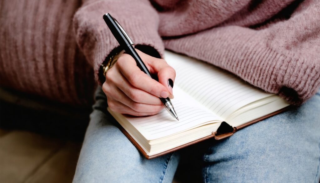 Close-up van vrouwelijke handen die in een notitieboek schrijven tijdens een coachingsessie, met een warme roze achtergrond.